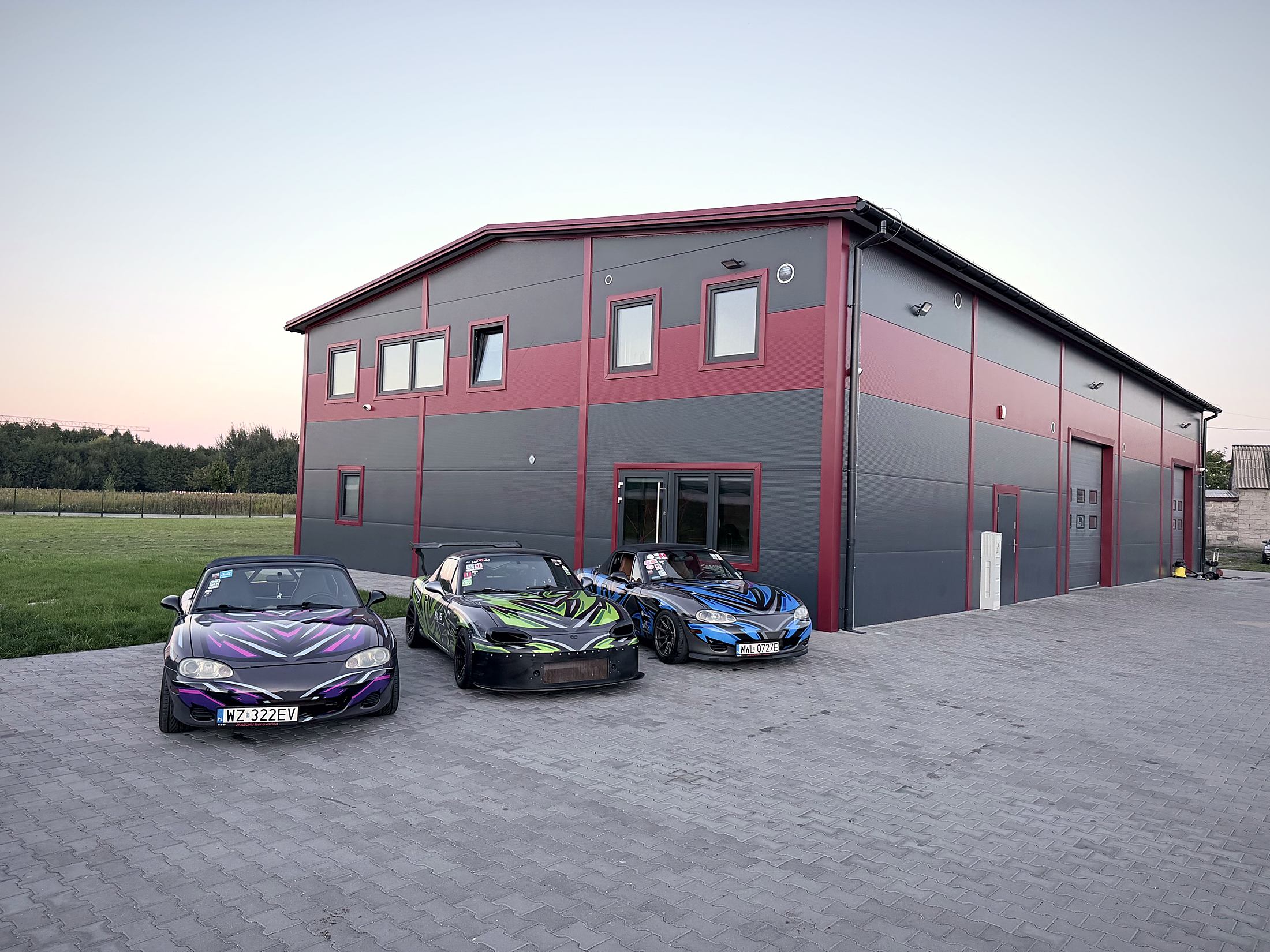 Budynek JKN Motors o zmierzchu, przed nim trzy Mazdy MX-5 w kolorowych oklejeniach